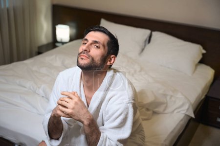 Photo for Sad man is sitting on a bed in a hotel room, he is in a comfortable bathrobe - Royalty Free Image