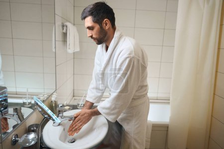 Téléchargez les photos : Homme d'âge moyen se lave les mains dans la salle de bain, il est dans un peignoir blanc - en image libre de droit