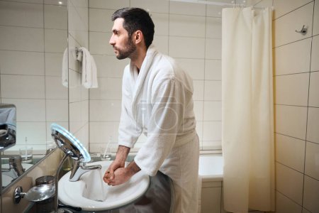 Foto de Hombre sin afeitar se lava las manos en el baño, mira su reflejo en el espejo - Imagen libre de derechos