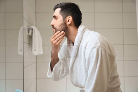 Photo for Man in a bathrobe looks in the mirror in the bathroom, male touches his unshaven face - Royalty Free Image