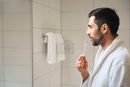 Téléchargez les photos : L'homme se tient devant un miroir dans la salle de bain, il a une brosse à dents dans les mains - en image libre de droit