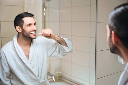Photo for Smiling middle-aged brunette brushes his teeth in front of a mirror, he is in a white bathrobe - Royalty Free Image
