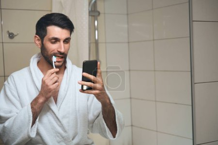 Téléchargez les photos : Brunette dans un peignoir se brosse les dents devant la caméra du téléphone, l'homme est situé devant un miroir - en image libre de droit