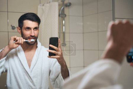Foto de Hombre guapo cepillándose los dientes delante de la cámara del teléfono, macho se encuentra delante del espejo - Imagen libre de derechos