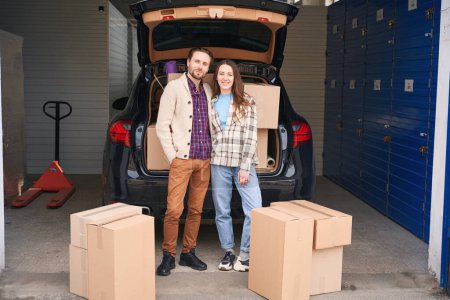 Photo for Smiling woman and man are standing near the open trunk of a car in a warehouse. Next to them are cardboard boxes - Royalty Free Image