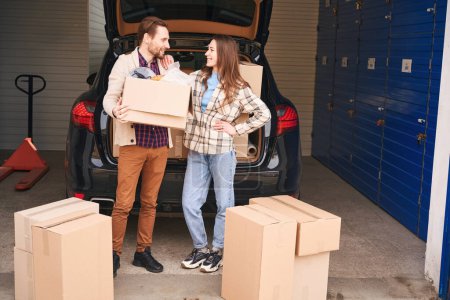 Photo for Smiling woman and man are standing near the open trunk of a car in a warehouse and looking at each other. Guy is holding a big cardboard box - Royalty Free Image