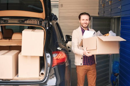 Photo for Happy man is standing near the open trunk of a car with boxes in a warehouse and holding a big cardboard box - Royalty Free Image