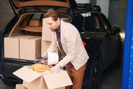 Photo for Guy is packing his things for moving into cardboard boxes in a storage warehouse. Nearby is a car with boxes in the trunk - Royalty Free Image