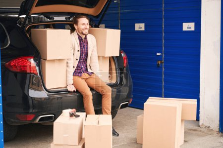 Photo for Young guy is sitting in the trunk of a car in a storage warehouse. Cardboard boxes with things for moving are in the trunk and next to the car - Royalty Free Image