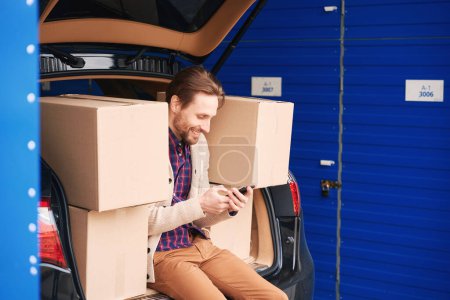Foto de El tipo feliz está sentado y llamando al maletero de un auto en un almacén. Cajas de cartón con cosas para mover están en el maletero - Imagen libre de derechos
