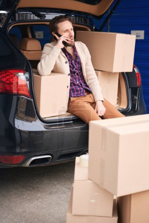 Photo for Pretty man is sitting and calling in the trunk of a car in a storage warehouse. Cardboard boxes with things for moving are in the trunk and next to the car - Royalty Free Image