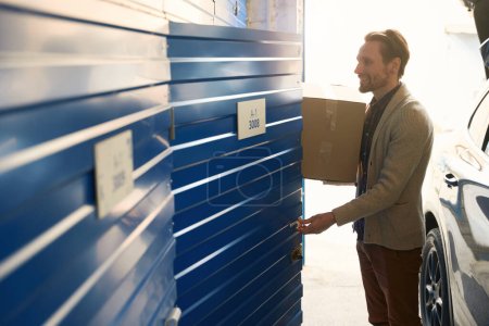 Téléchargez les photos : Un gars souriant ouvre la salle de stockage dans l'entrepôt et tient une grande boîte en carton - en image libre de droit