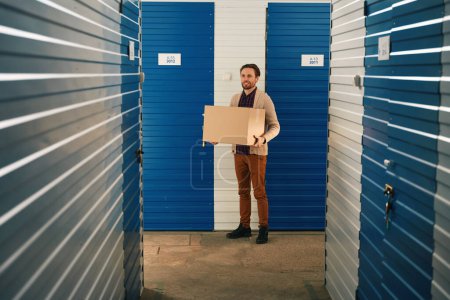 Photo for Happy guy is holding a big cardboard box and standing in storage warehouse - Royalty Free Image