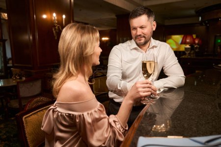 Téléchargez les photos : Heureux couple avec des verres de vin mousseux avoir la conversation tout en étant assis au comptoir du bar - en image libre de droit