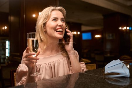 Téléchargez les photos : Souriant belle femme parlant au téléphone et buvant du vin mousseux tout en étant assis au comptoir du bar - en image libre de droit