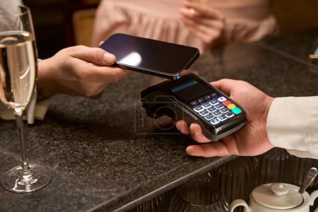 Photo for Close up of man making payment using telephone by nfc technology with his woman while barmen holding credit card reader at bar counter - Royalty Free Image