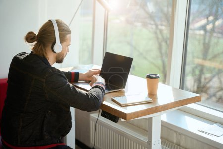 Foto de Hombre guapo con corte de pelo elegante que trabaja en el ordenador portátil mientras está sentado en la ventana en la oficina - Imagen libre de derechos