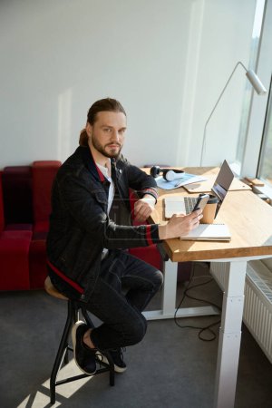 Photo for Handsome man looking at camera while using cell phone and sitting on chair at at coworking space - Royalty Free Image