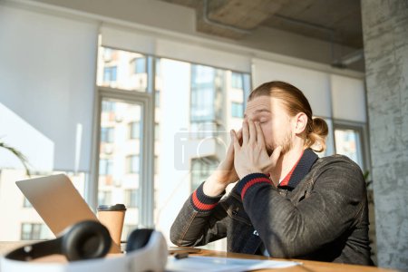 Téléchargez les photos : Jeune homme fermé les yeux tout en travaillant sur ordinateur portable et assis près de la fenêtre à l'intérieur - en image libre de droit