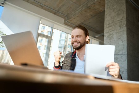 Téléchargez les photos : Travailleur masculin avec du papier à la main heureux tout en faisant des tâches sur ordinateur portable à l'intérieur - en image libre de droit