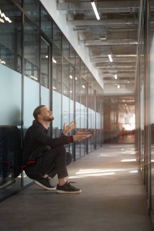 Téléchargez les photos : Homme élégant avec une coupe de cheveux élégante levant les yeux tout en étant assis sur ses hanches dans le couloir du bureau - en image libre de droit