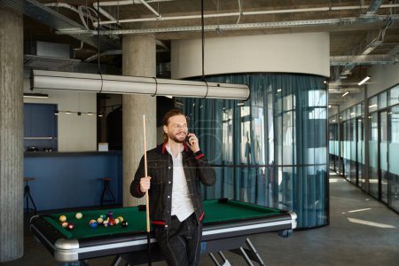 Photo for Attractive male with happy facial expression standing near pool table while having dialogue by cell phone indoors - Royalty Free Image