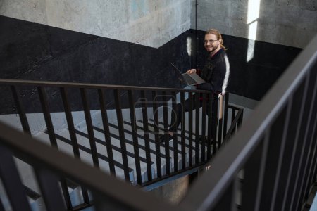 Foto de Hombre diligente con computadora portátil en sus manos mirando a la cámara y sonriendo mientras subía las escaleras - Imagen libre de derechos