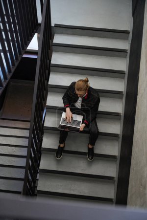 Photo for Top view on concentrated man with stylish haircut typing on laptop while sitting on stairs - Royalty Free Image