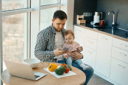 Photo for Male in home clothes sits at the kitchen table, he has a cute baby in his arms - Royalty Free Image