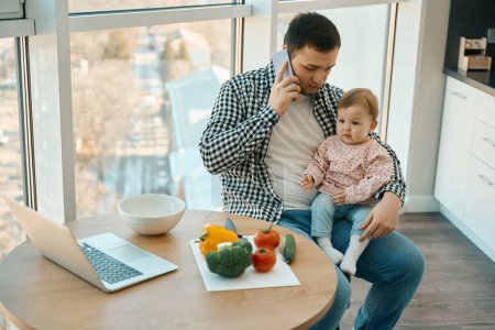 Téléchargez les photos : L'homme parle au téléphone avec un bébé dans ses bras, un ordinateur portable et des légumes sont sur la table de cuisine - en image libre de droit