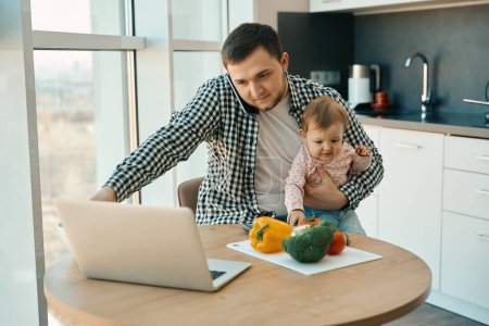 Téléchargez les photos : Homme barbu travaille de la maison avec un bébé dans ses bras, ordinateur portable et une variété de légumes sont sur la table - en image libre de droit