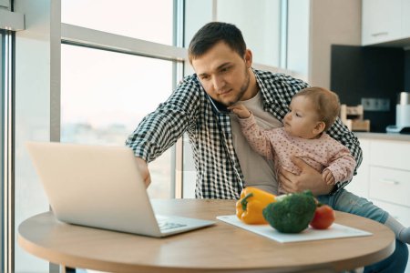 Téléchargez les photos : Homme barbu travaille de la maison avec un enfant dans ses bras, ordinateur portable et une variété de légumes sont sur la table - en image libre de droit