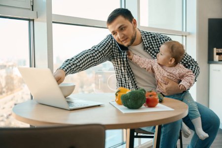 Téléchargez les photos : Père aimant travaille de la maison avec un enfant dans ses bras, ordinateur portable et une variété de légumes sur la table - en image libre de droit