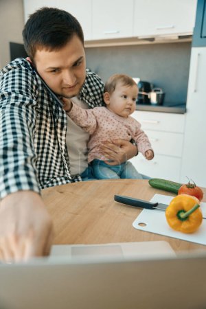 Photo for Young dad works from home on a laptop, he has a baby daughter in his arms - Royalty Free Image