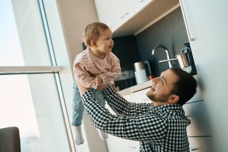 Photo for Happy dad and baby daughter joyfully spend time at home in the kitchen, the man raised the child high up - Royalty Free Image