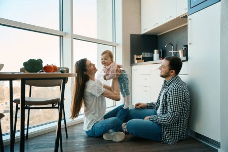 Téléchargez les photos : Les parents aidants jouent avec un petit enfant à la maison dans la cuisine, la famille est située à la fenêtre de vue - en image libre de droit