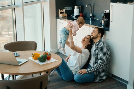 Téléchargez les photos : Joyeuses maman et papa jouent avec le bébé à la maison, la famille est située sur le sol dans la cuisine - en image libre de droit
