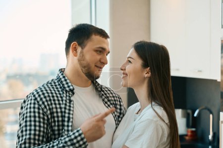 Téléchargez les photos : Les jeunes époux souriants sont debout, embrassant, à la fenêtre de la vue dans la cuisine, les gars en vêtements confortables décontractés - en image libre de droit