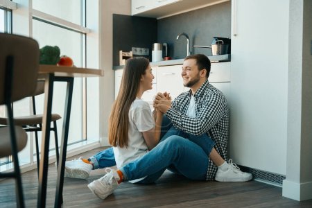 Photo for Smiling man and woman sit hugging on the floor in the kitchen, they gently hold hands - Royalty Free Image