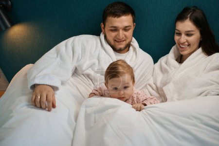 Photo for Happy young parents admiring their pretty baby, the family settled down on a big bed - Royalty Free Image