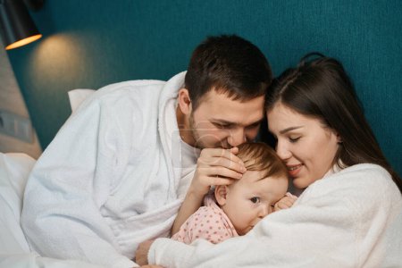 Téléchargez les photos : Bébé mignon dans les bras des mamans dans le lit des parents, papa embrasse l'enfant sur le dessus de sa tête - en image libre de droit