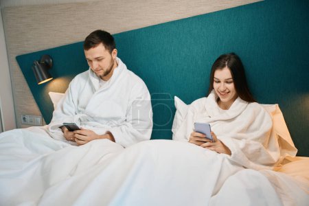 Téléchargez les photos : Les jeunes conjoints se reposent dans un lit doux dans des vêtements confortables à la maison, ils ont des téléphones mobiles dans leurs mains - en image libre de droit