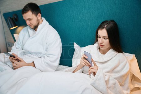 Téléchargez les photos : L'homme et une femme se reposent dans un lit doux dans des vêtements confortables à la maison, ils ont des téléphones mobiles dans leurs mains - en image libre de droit
