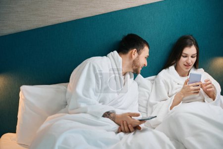 Téléchargez les photos : Homme souriant regardant son téléphone femme, couple relaxant sur lit doux - en image libre de droit