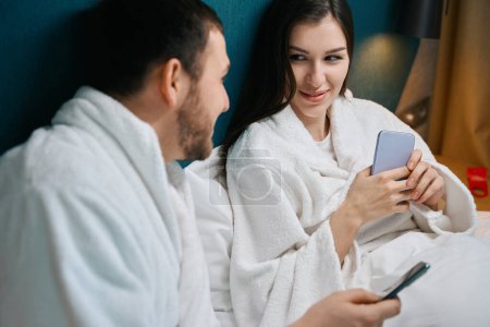 Téléchargez les photos : L'homme et une femme se prélassent dans un lit doux avec des téléphones, les gars sont de bonne humeur - en image libre de droit