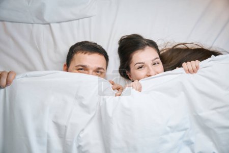 Téléchargez les photos : Souriant mari et femme se prélasser sous les couvertures sur un grand lit, ils ont l'air mignon sous les couvertures - en image libre de droit