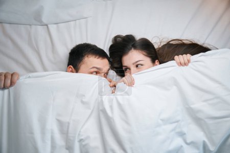 Téléchargez les photos : Mari mignon et femme se prélassent sous les couvertures sur un grand lit, ils se regardent drôlement sous les couvertures - en image libre de droit