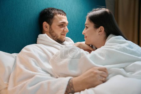 Photo for Smiling charming woman looks tenderly at her husband, the spouses bask under the covers on a large bed - Royalty Free Image