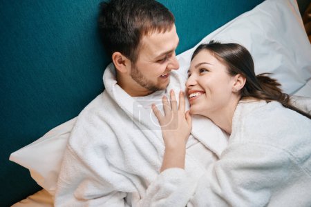 Téléchargez les photos : Femme amoureuse regarde son mari avec tendresse, les conjoints se prélassent sous les couvertures sur un grand lit - en image libre de droit