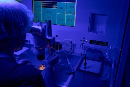 Foto de Embriólogo mirando en microscopio, realizando cultivo de embriones, comprobando indicadores de salud en el monitor, trabajando en luz ultravioleta - Imagen libre de derechos